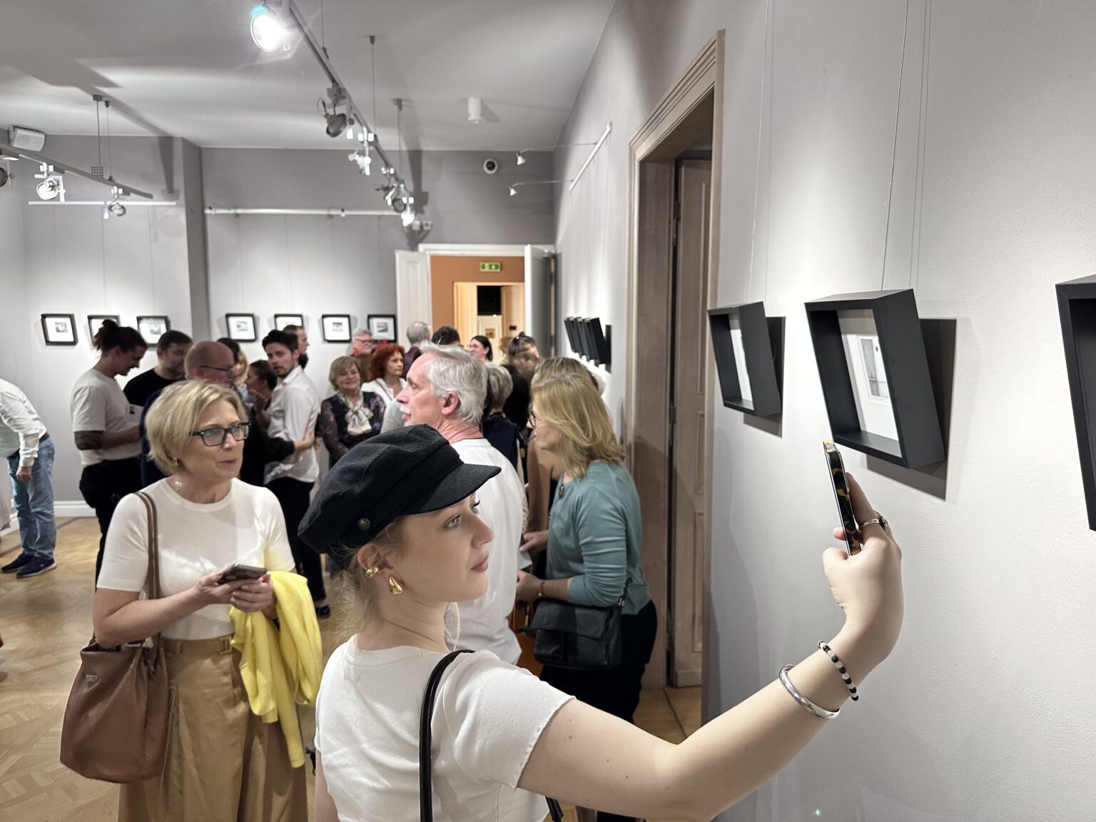 Grpa zwiedzających wystawę. Na pierwszym planie kobieta w czapce robi zdjęcie smartfonem