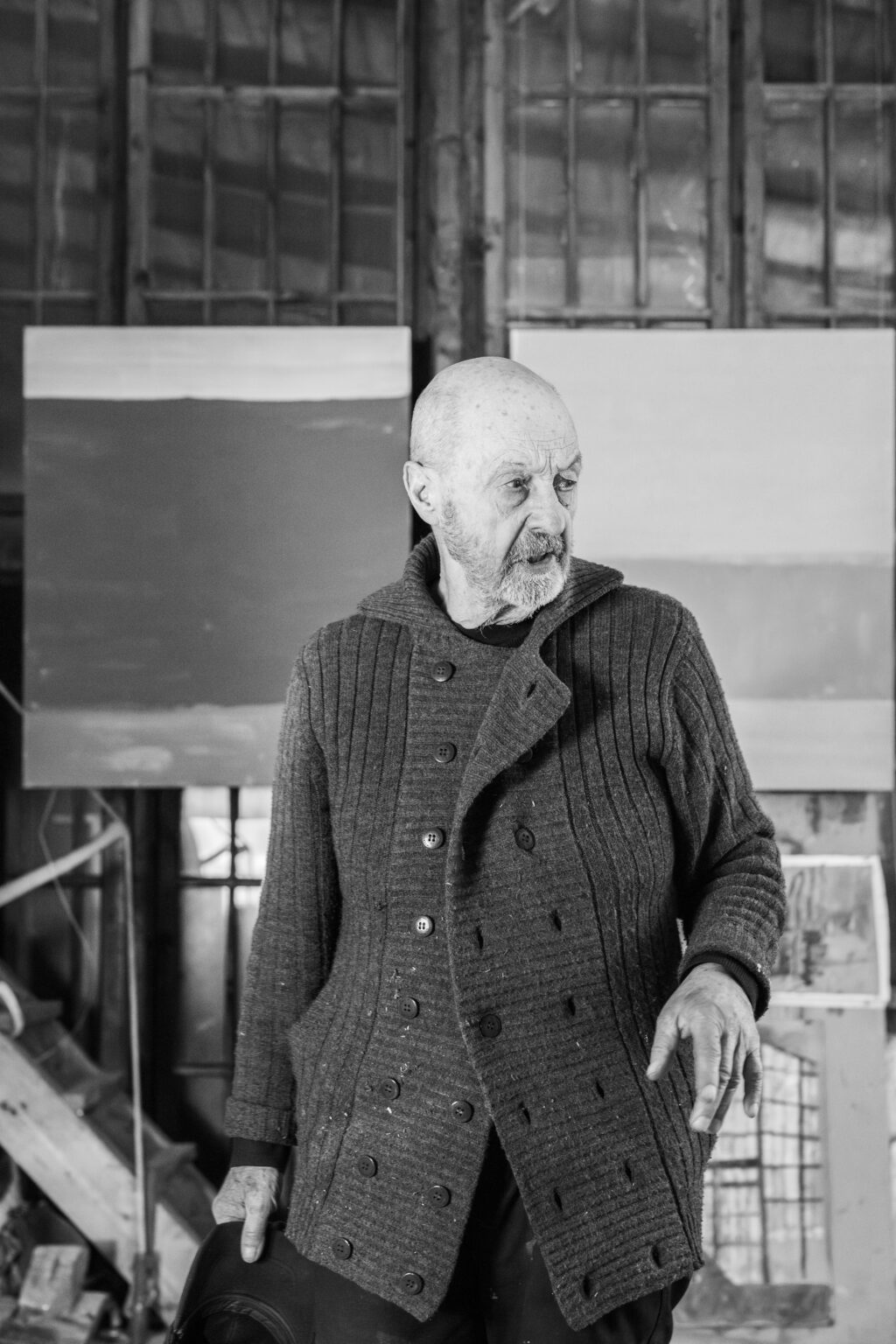 Czarno-biały portret mężczyzny w grubym swetrze zapinanym na dwa rzędy guzików. Obrazy w tle