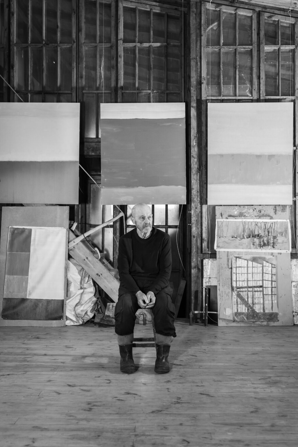 Czarno-białe zdjęcie mężczyzny w gumiakach, siedzącego na stołku. W tle obrazy.