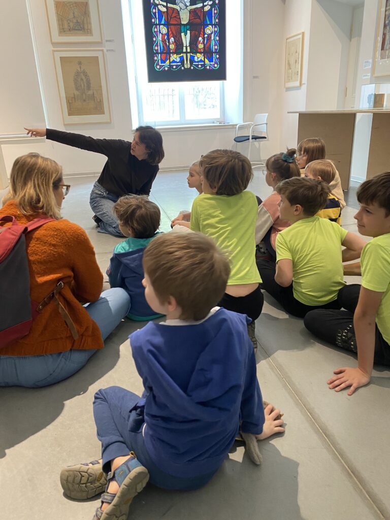 Grupa dzieci siedzi na podłodze, z nimi prowadząca, pokazuje coś po lewej stronie. Wszyscy patrzą w tamtym kierunku