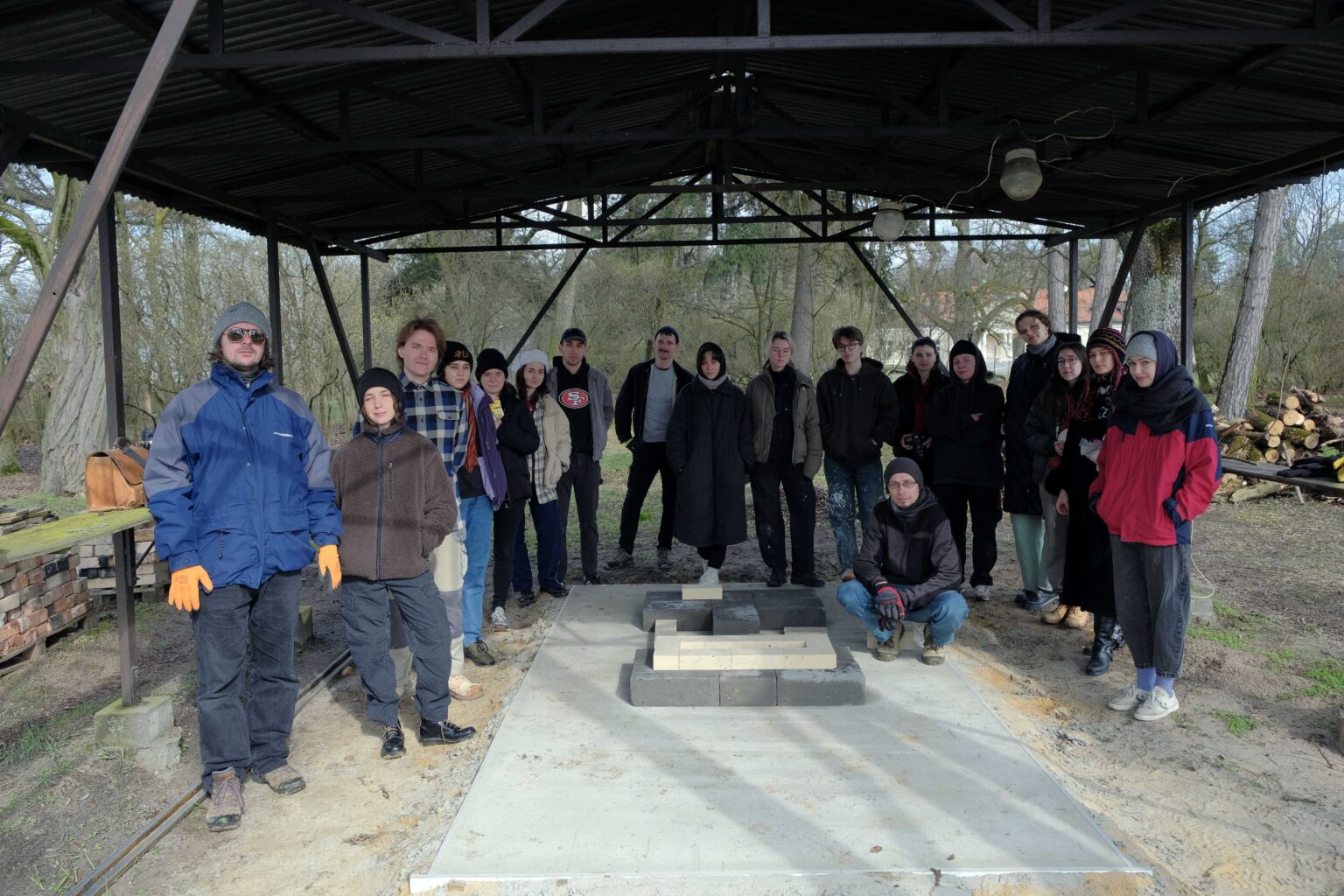 Grupa osób na zewnątrz pod dachem, wokół miejsca wyłożonego cegłami