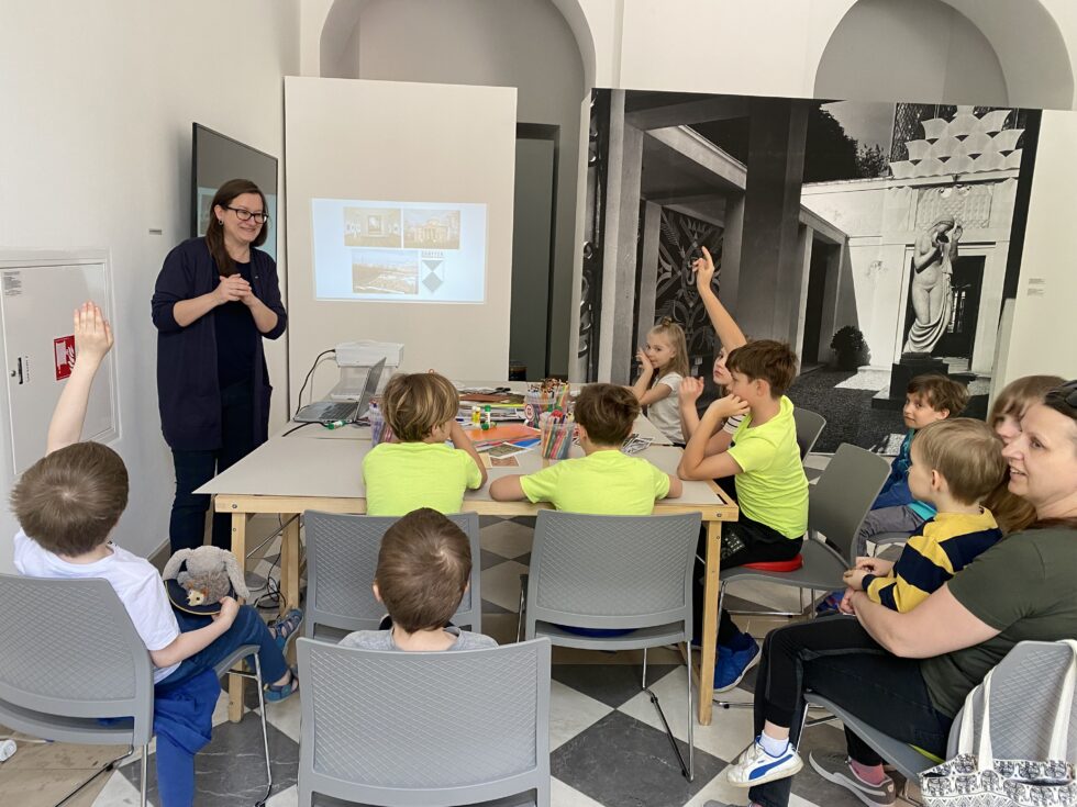 Grupa dzieci na warsztatach. Po prawej, przy stole stoi prowadząca,