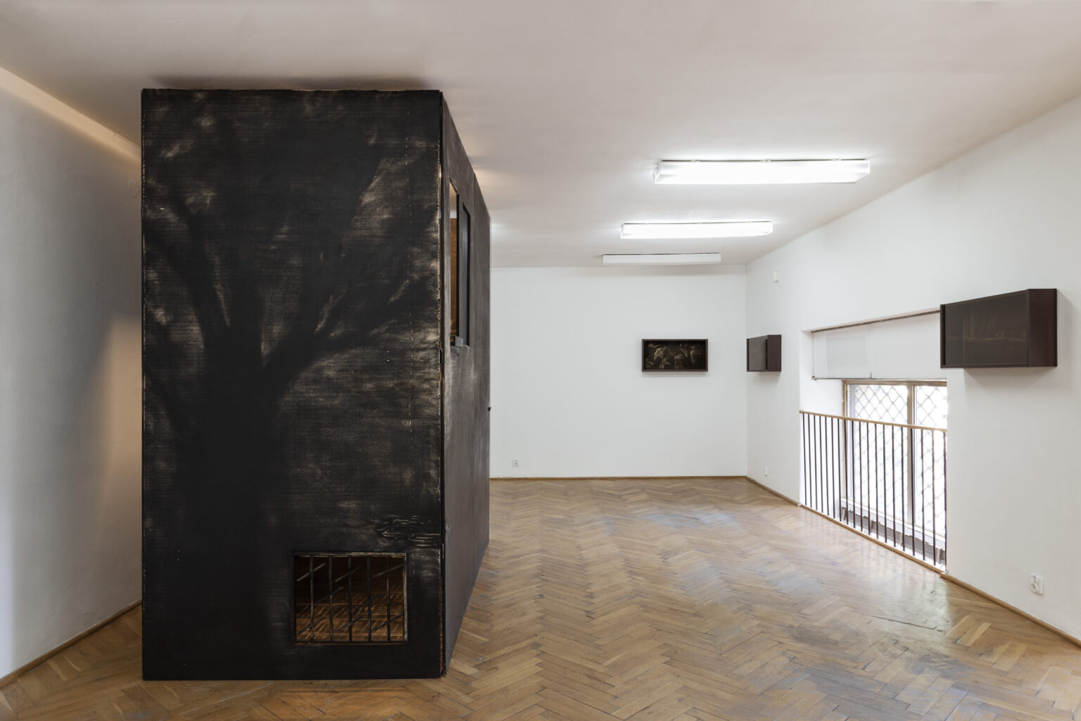 Duży czarny obiekt zajmujący przestrzeń od podłogi do sufitu jasnego pomieszczenia z jarzeniówkami i drewnianą klepką. trzy mniejsze czarne obiekty na ścianach.