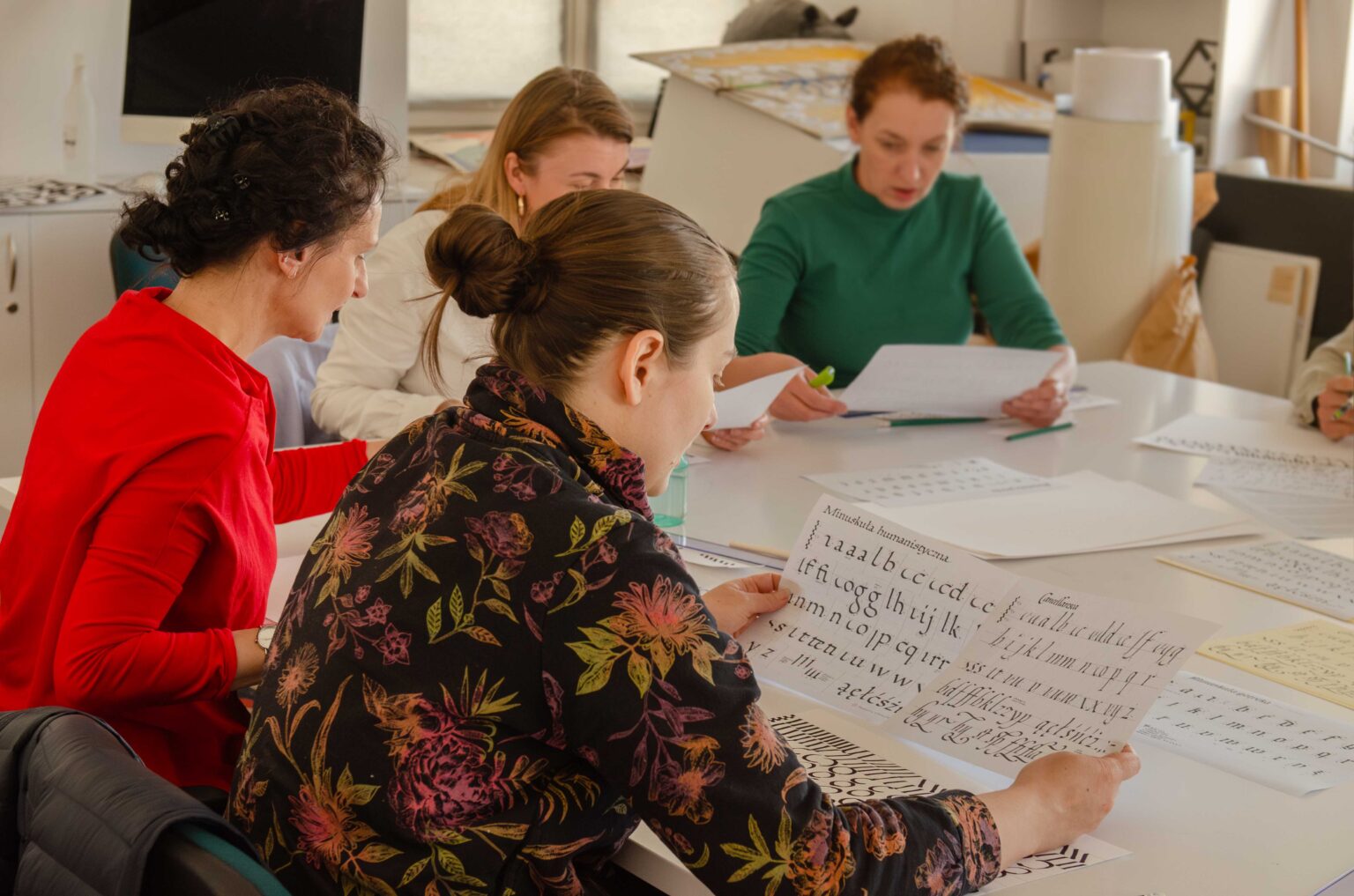 cztery osoby przy stole w pracowni; jedna z nich trzyma w dłoniach arkusz papieru z wykaligrafowanymi literami