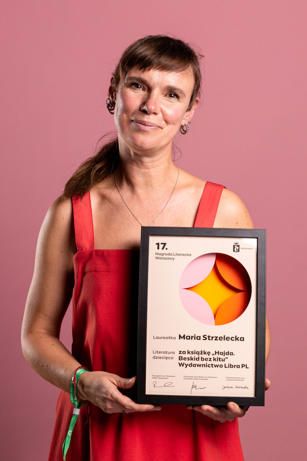 Portret uśmiechniętej kobiety w pomarańczowej sukience, prezentującej dyplom.