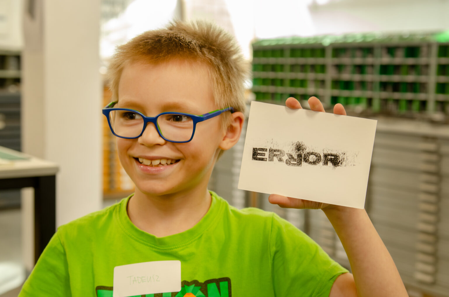 Uśmiechnięty chłopiec w okulrach trzyma w ręku podłużną kartkę z grafiką