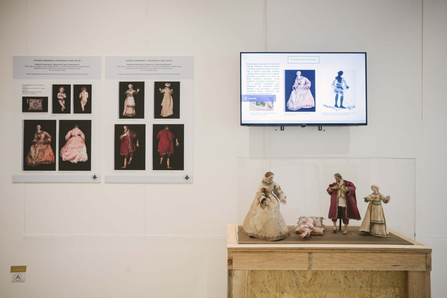 Widok wystawy: fragment wydrukw barwnych na ścianie, monitor, a pod nim wyeksponowane figurki