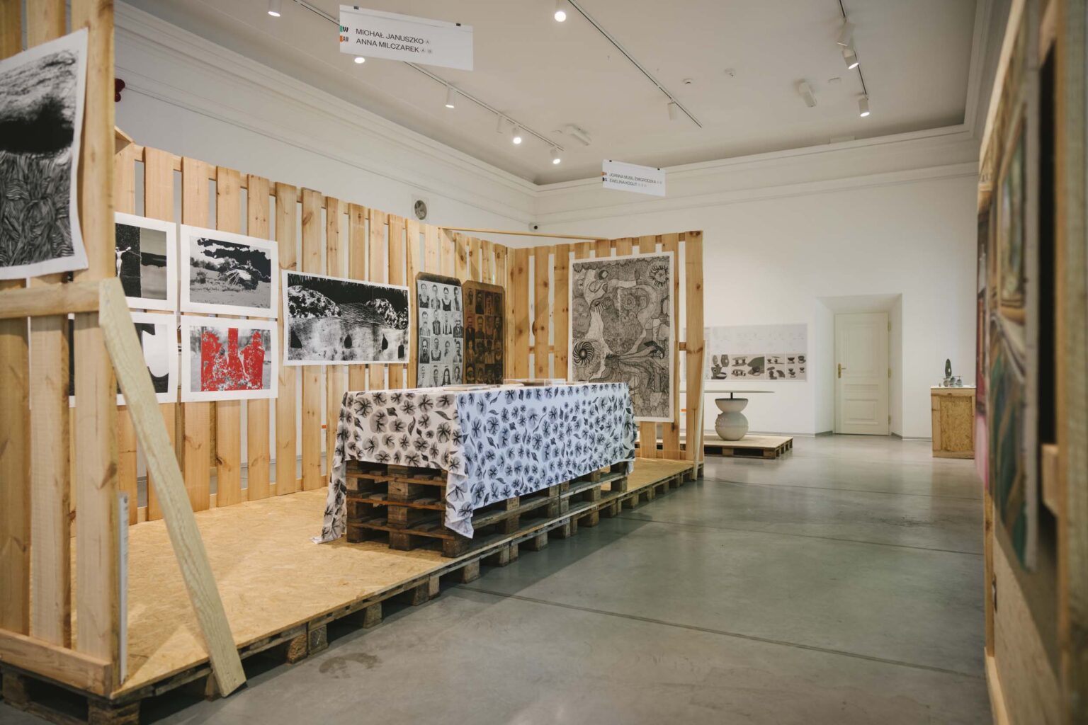 widok wystawy, w drewnianej instalacji, imitującej przekrój pomieszczenia imitacja społu przykrytego obrusem, sitodruki na "ścianie"