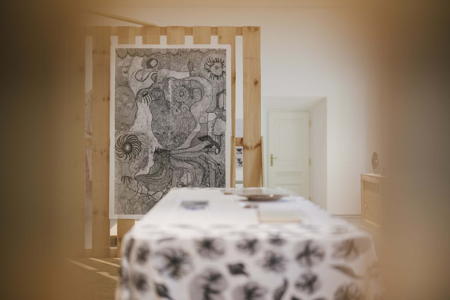 Na pierwszym planie stół nakryty białym obrusem we florystyczne, czarno-białe motywy, w tle czarno-biały sitodruk