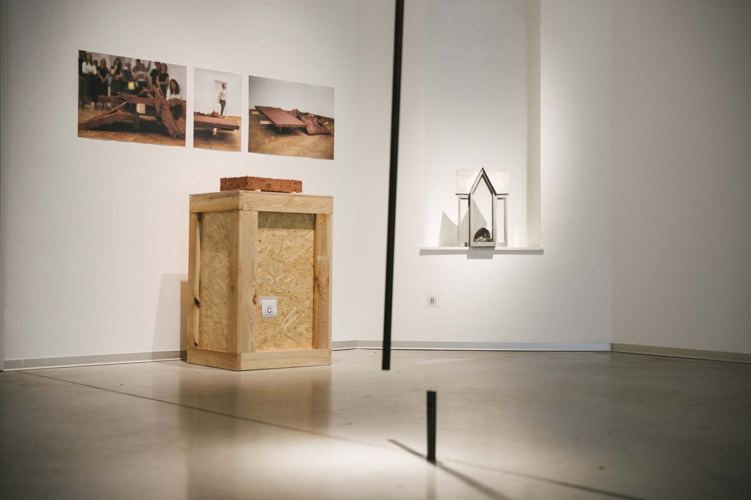 Pionowa instalacja złożona z krótkiej części rurki przytwierdzonej do podłogi oraz długiej, ruchomej, przytwierdzonej do sufitu. W tle: zdjęcia na ścianie, kadzielnica we wnęce ściany i obiekt gliniany na drewnianym postumencie