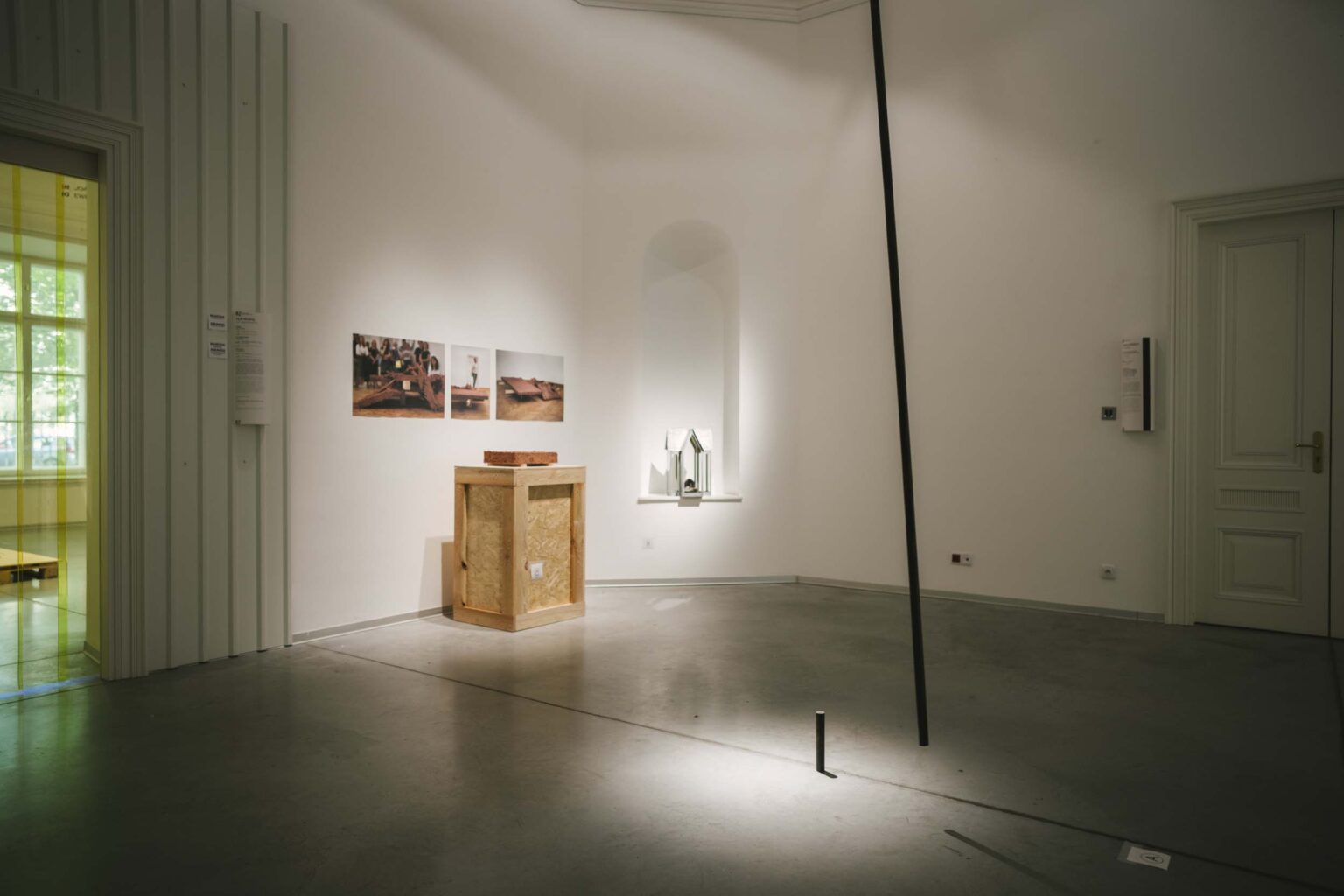 Pionowa instalacja złożona z krótkiej części rurki przytwierdzonej do podłogi oraz długiej, ruchomej, przytwierdzonej do sufitu. W tle: zdjęcia na ścianie, kadzielnica we wnęce ściany i obiekt gliniany na drewnianym postumencie