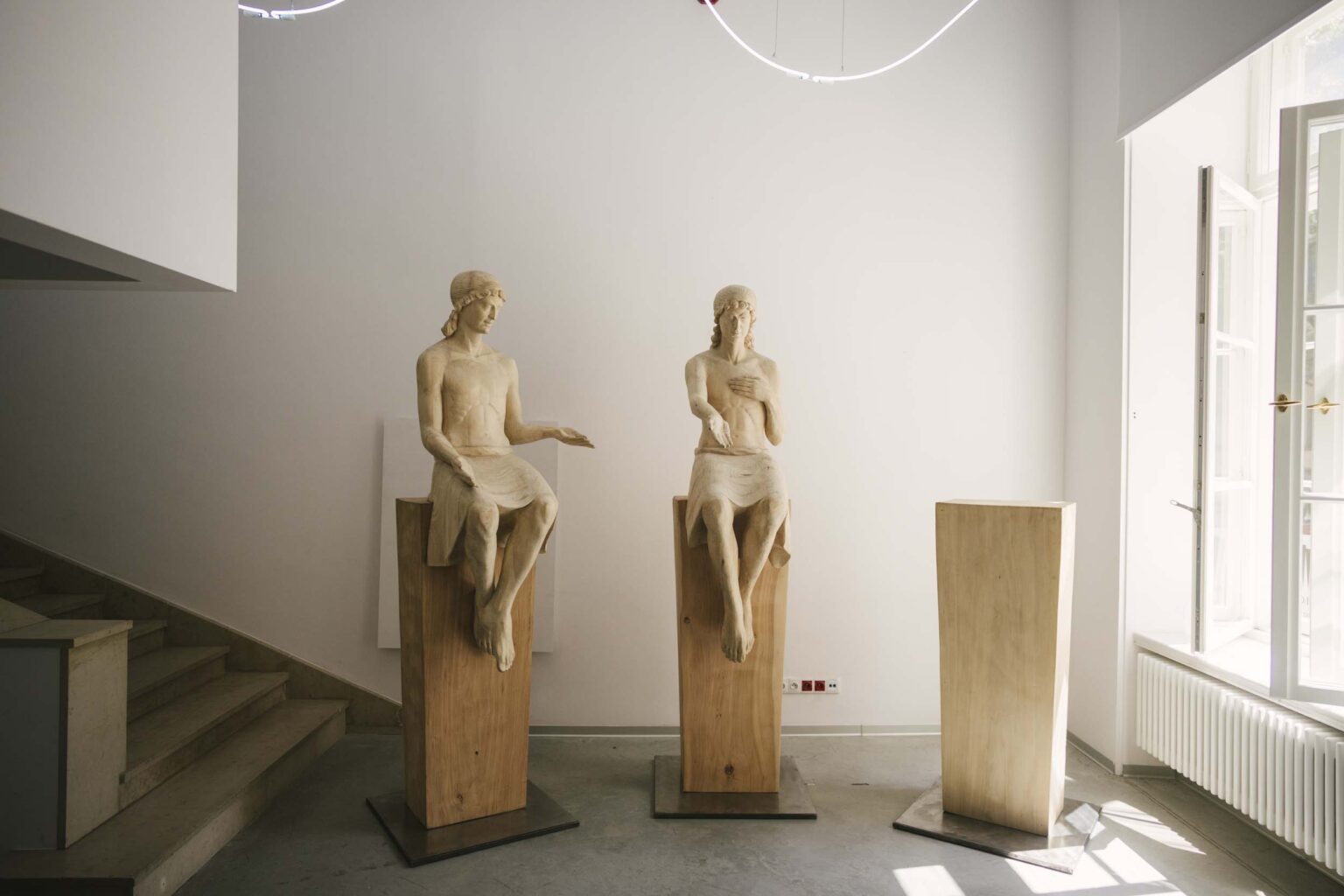 Dwie naturalnej wielkości drewniane rzeźby przedstawiające osoby siedzące na postumentach