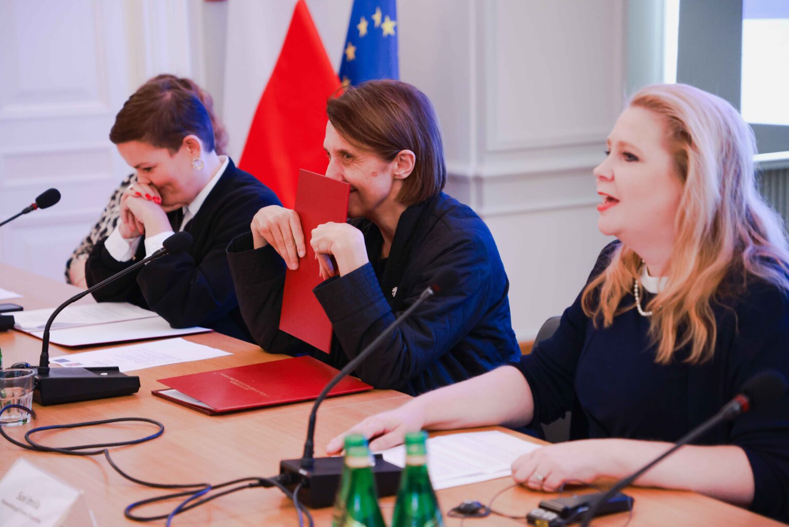 trzy kobiety za biurkiem konferencyjnym, w tle flagi: polski i UE