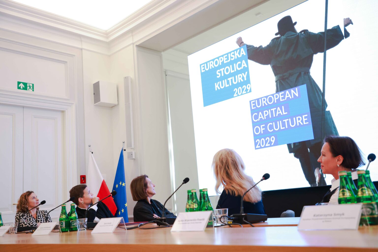 trzy kobiety za biurkiem konferencyjnym, odwrócone do tyłu patrzą na ekran