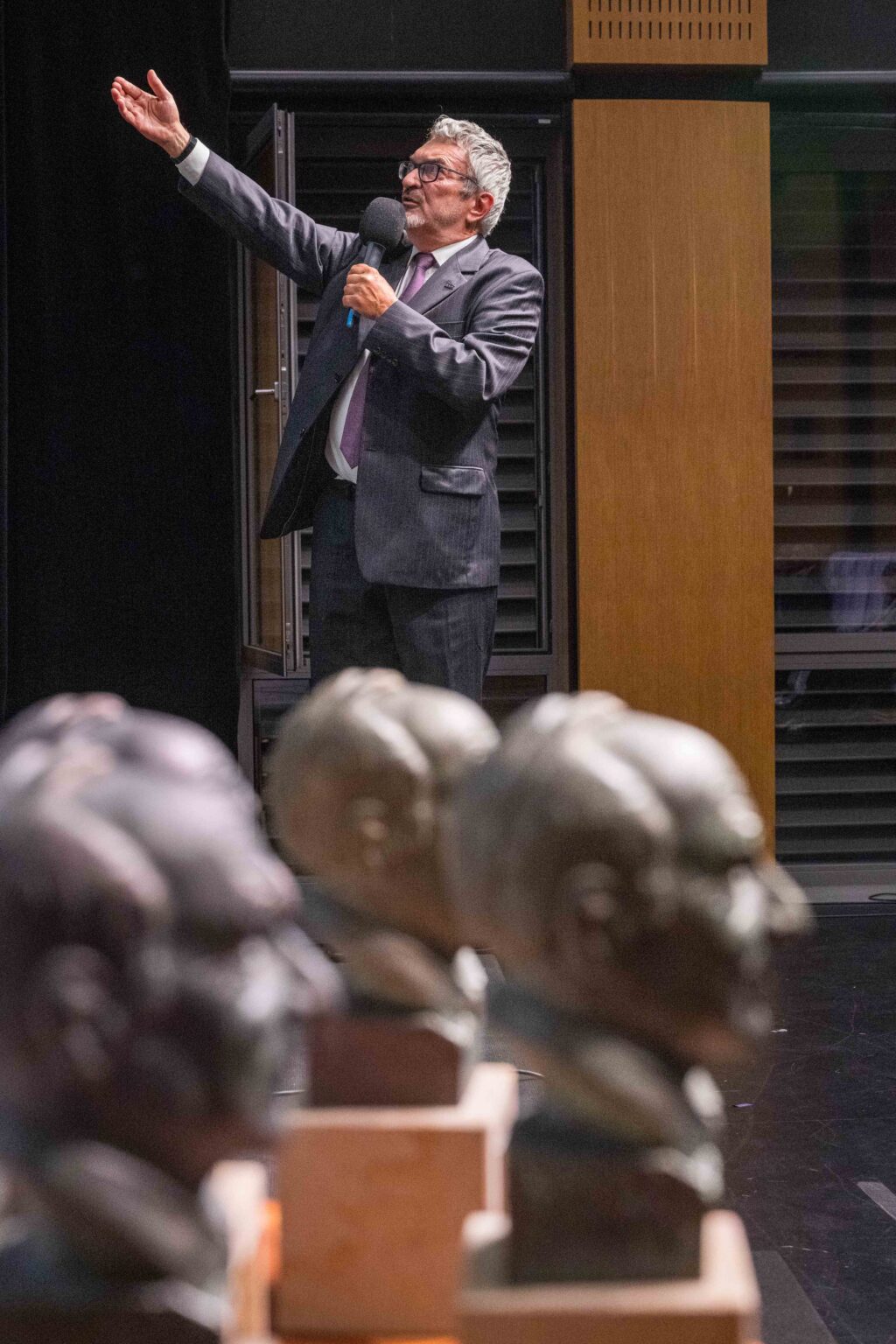 mężczyzna w garniturze mówi do mikrofonu, unosi rękę wysoko; na pierwszym planie 3 statuetki