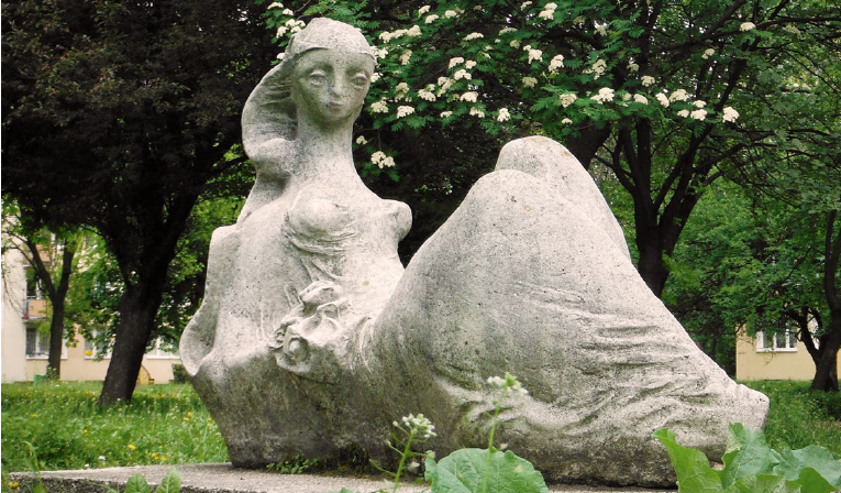 Kolorowe zdjęcie białej rzeźby przedstawiającej siedzącą kobietę, w tle kwitnące drzewa
