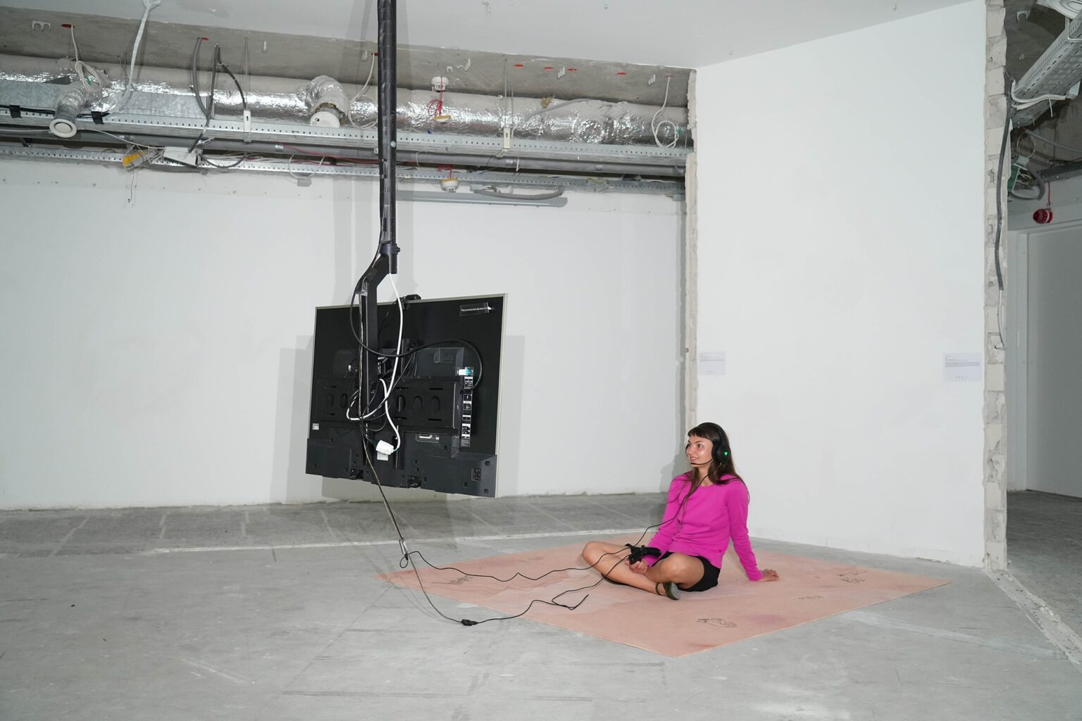 kobita w różowym swetrze siedzi na podłodze i patrzy na podwieszony z sufitu monitor; surowe wnętrze