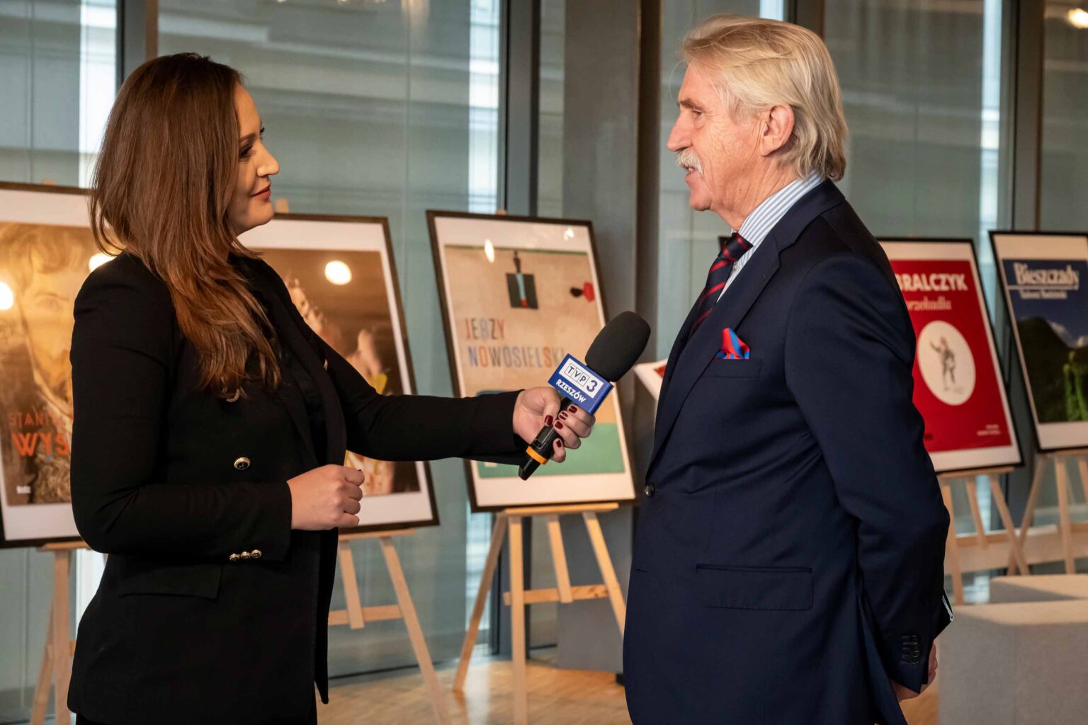 Mężczyzna w garniturze udziela wywiadu kobiecie z mikrofonem, plakaty na sztalugach w tle