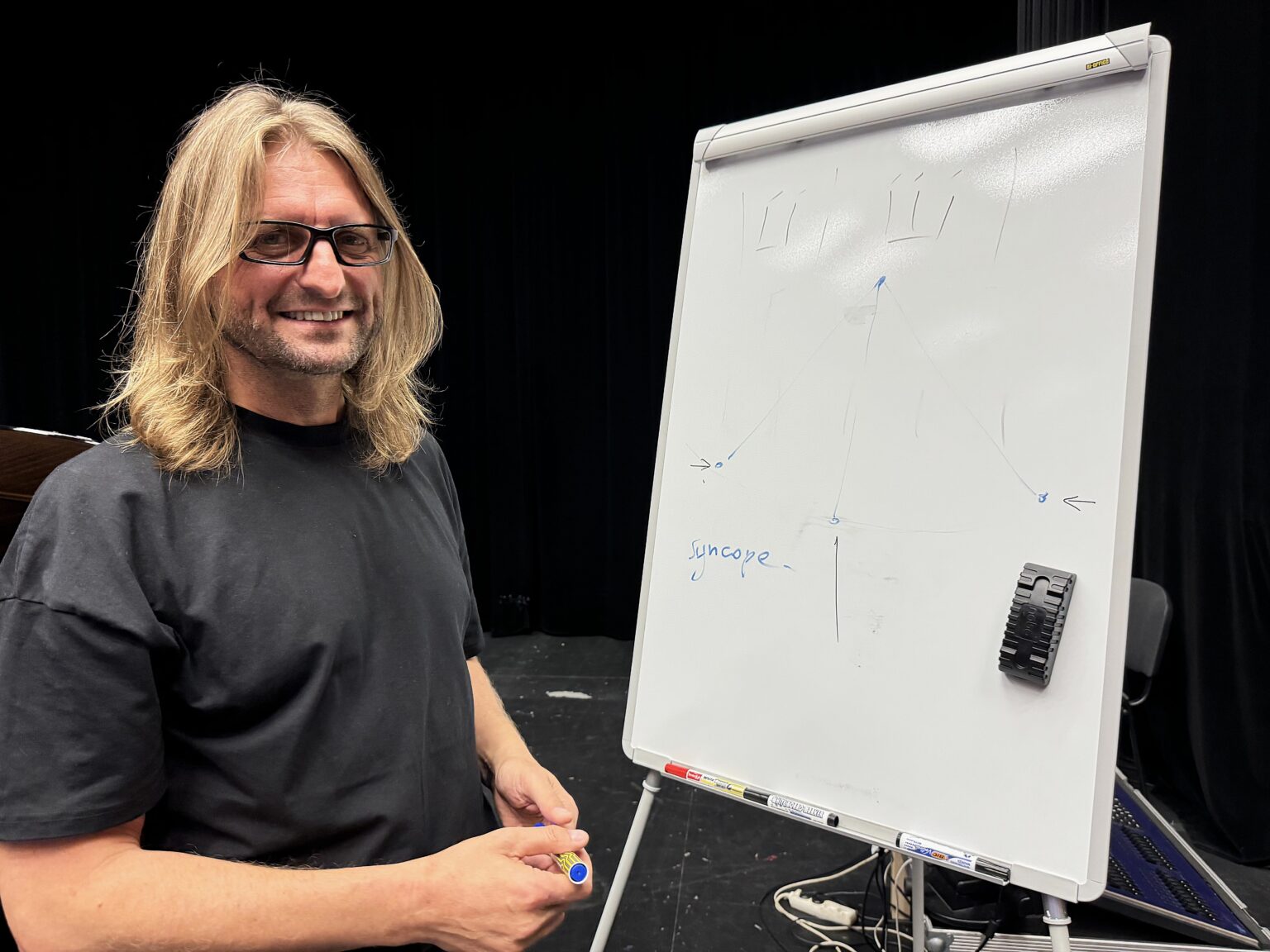 Portret uśmiechniętego, długowłosego mężczyzny w okularach przy tablicy.