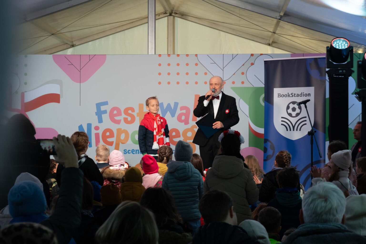 mężczyzna w garniturze i chłopiec na scenie. Przed sceną duża grupa dzieci.