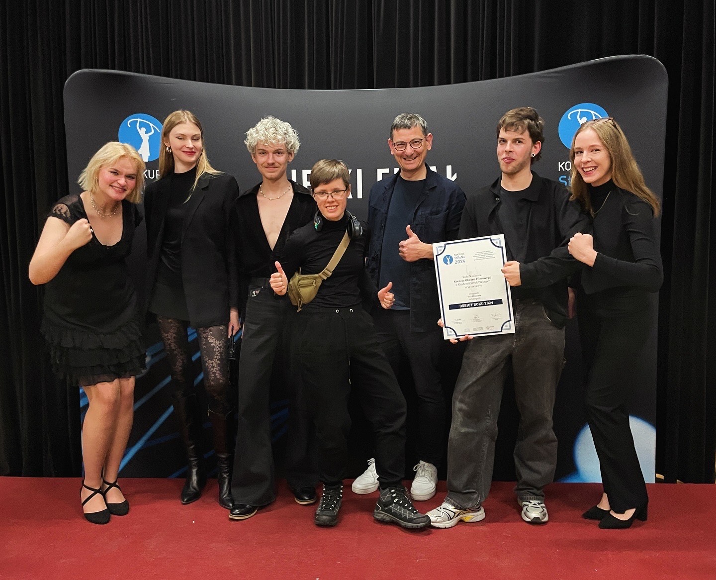 Grupa roześmianych osób na scenie prezentuje dyplom