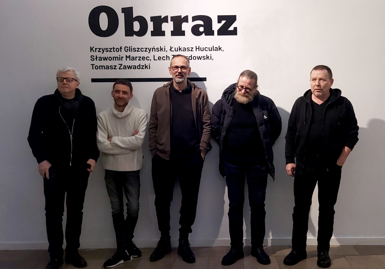 5 mężczyzn stoi w rzędzie, za nimi na ścianie napis: "Obrraz"