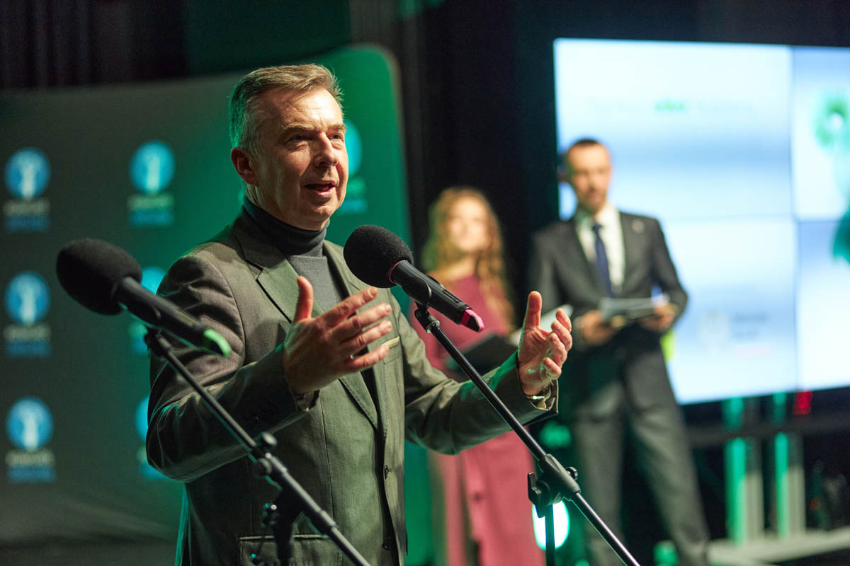 mężczyzna w garniturze przy mikrofonie z rękami w geście powitania gości