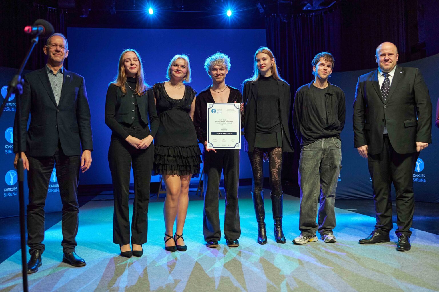 grupa osób na scenie w rzędzie, osoba w środku trzyma dyplom