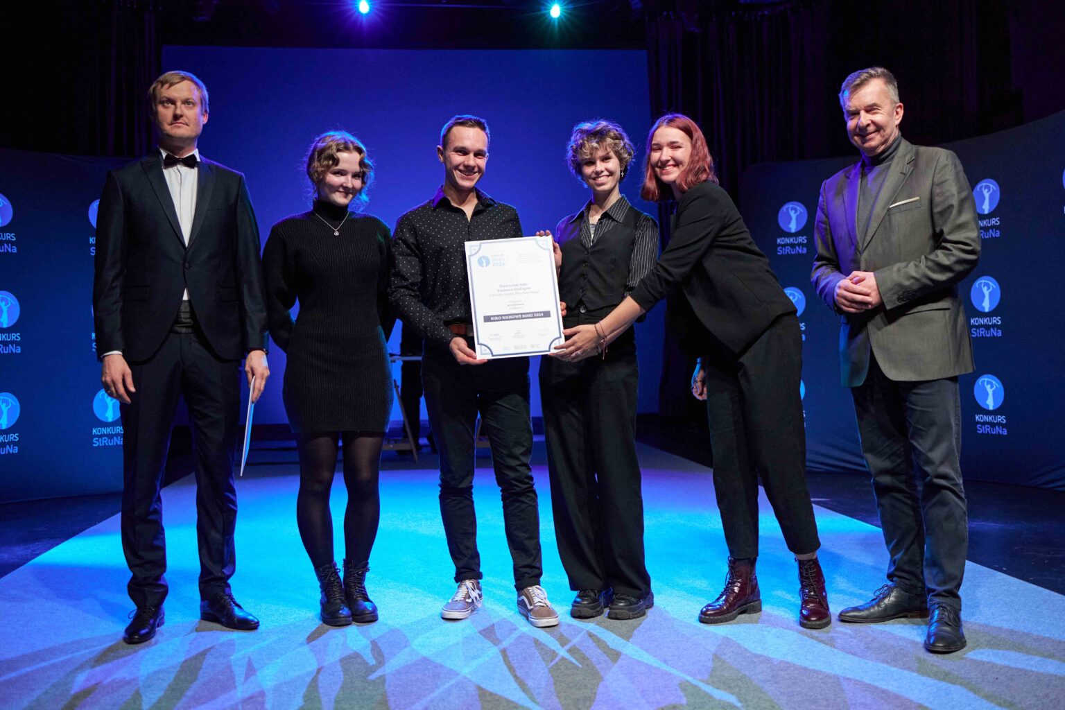 grupa osób stoi na scenie w rzędzie, osoba w środku trzyma dyplom