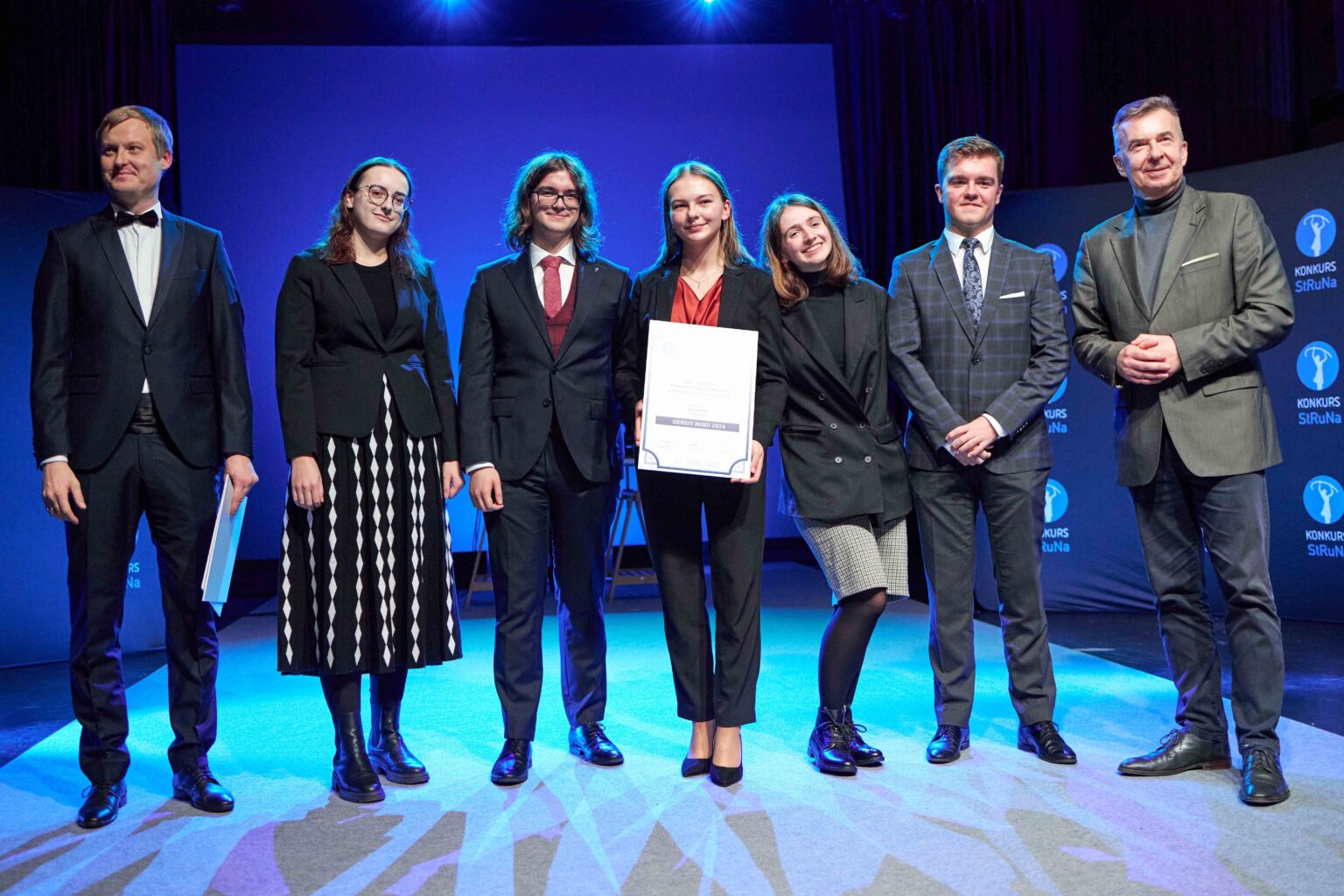 grupa osób stoi na scenie w rzędzie, osoba w środku trzyma dyplom