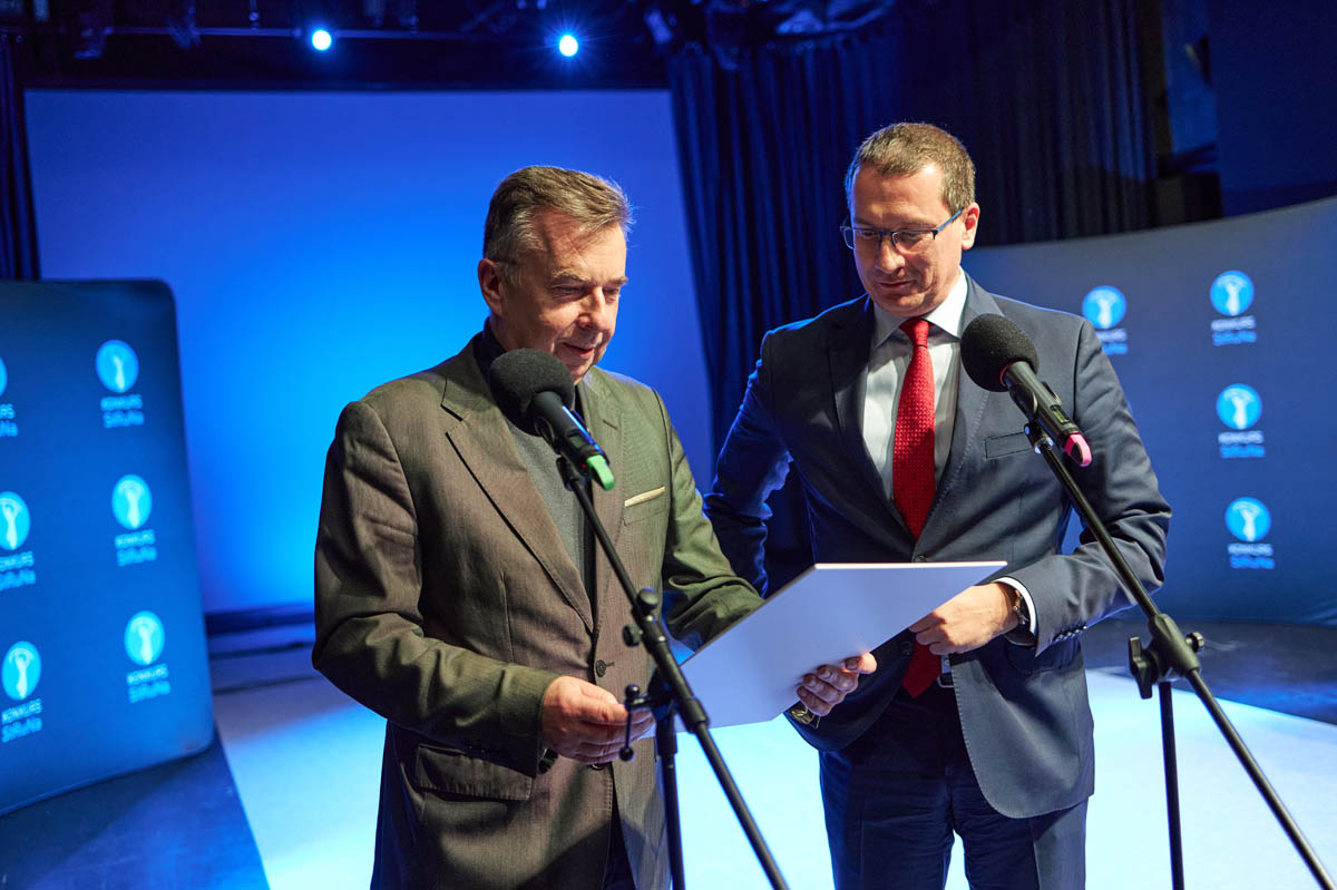 dwaj mężczyźni na scenie przy mikrofonach, jeden z nich odczytuje coś z kartki