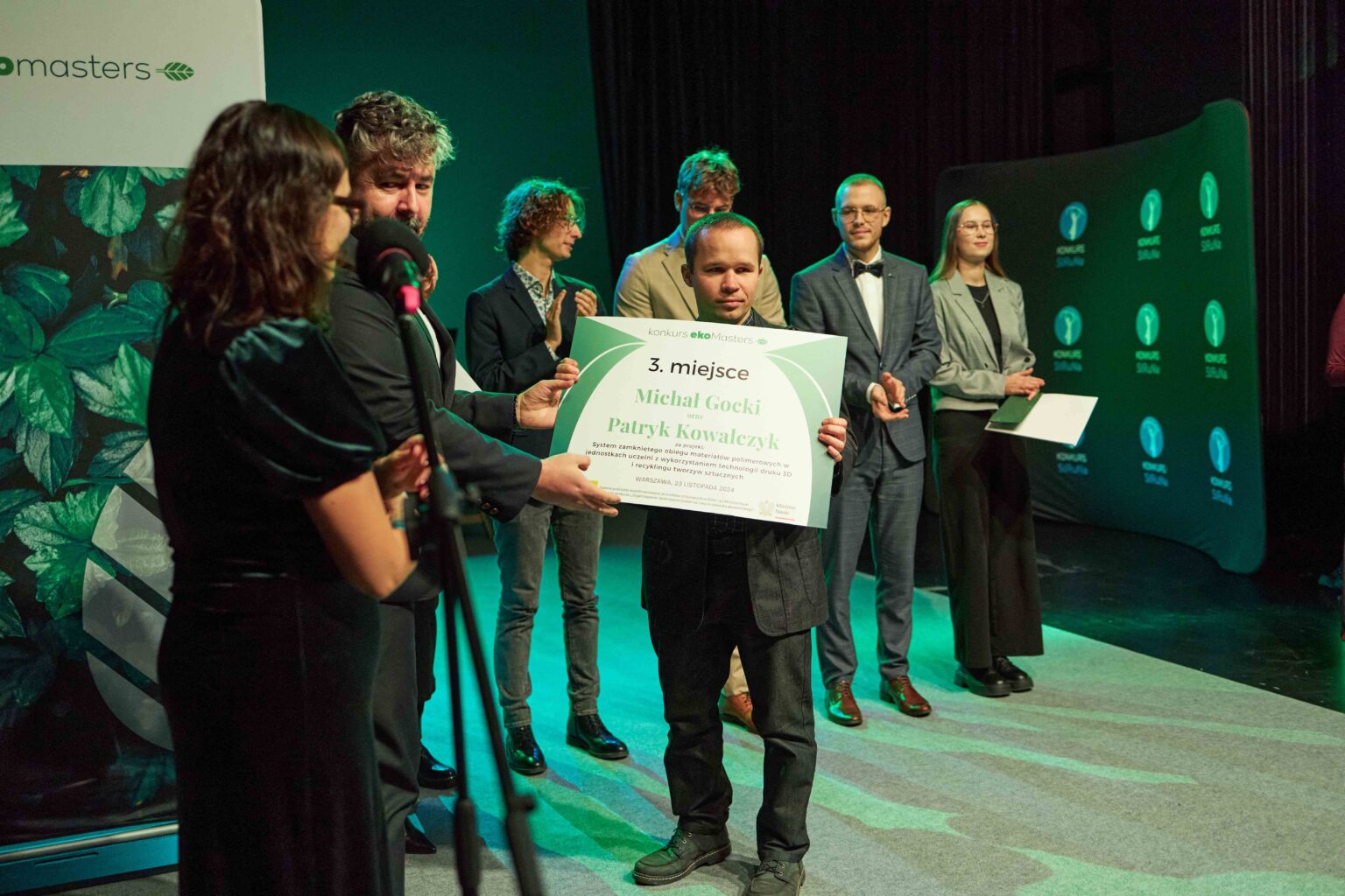 mężczyzna na scenie z dyplomem, grupa osób wokół