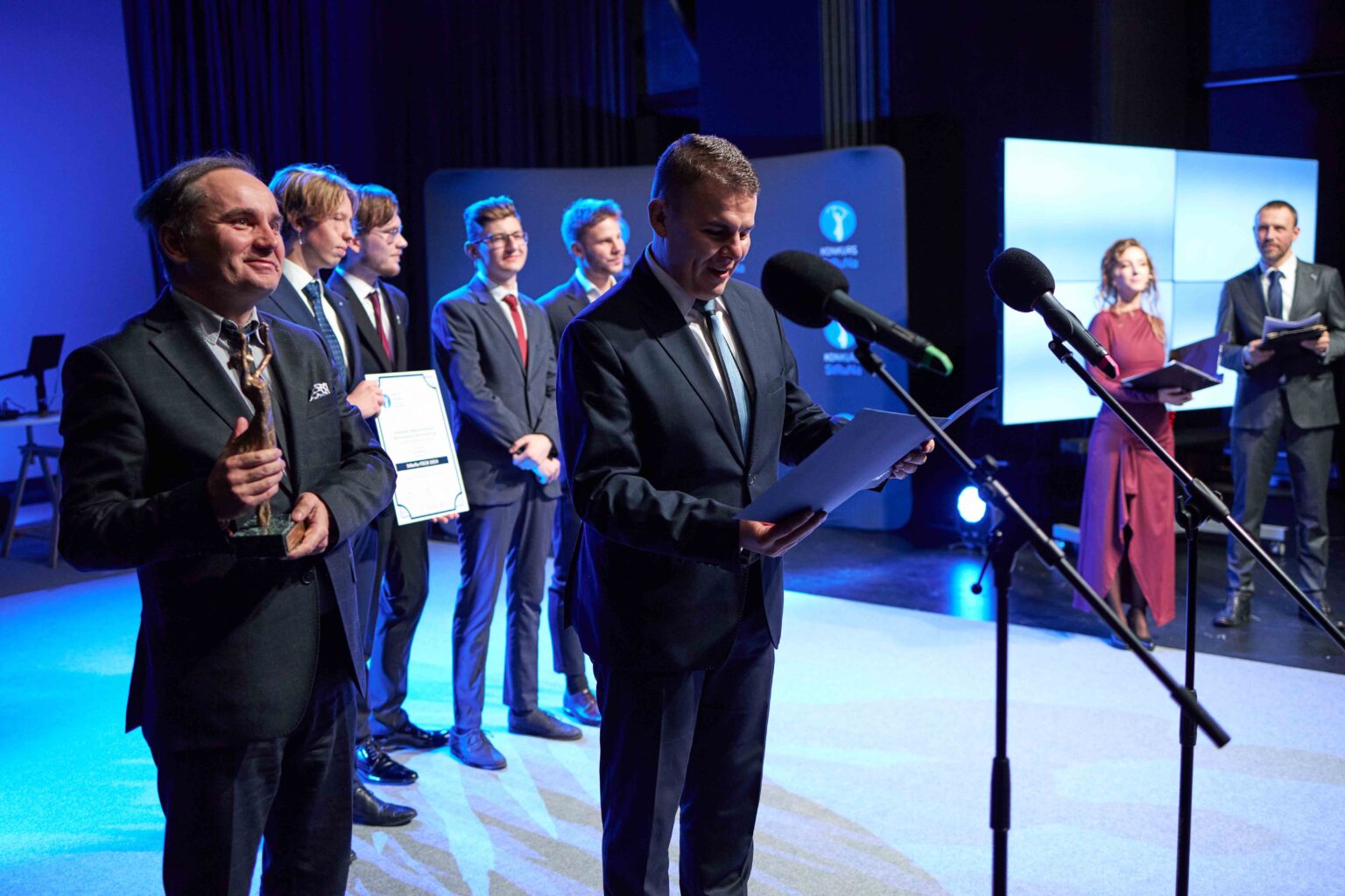 grupa osób na scenie; mężczyzna przy mikrofonie