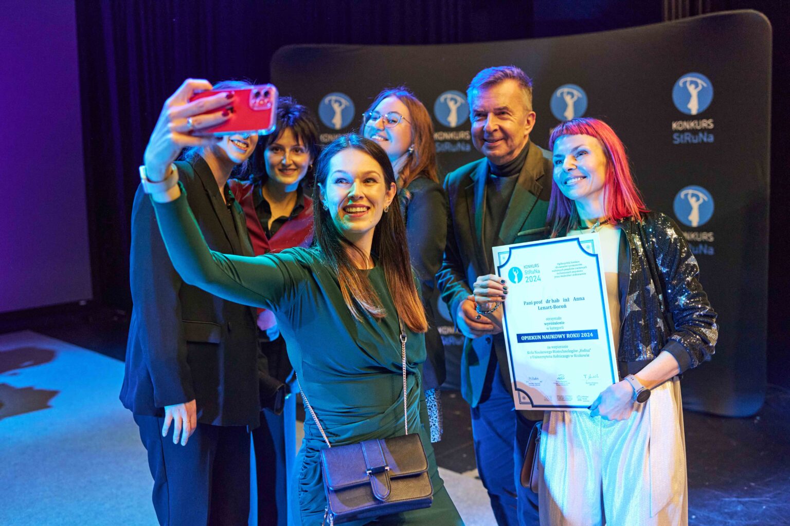 grupa osób na scenie, dziewczyna z przodu robi selfie sobie i osobom stojącym za nią