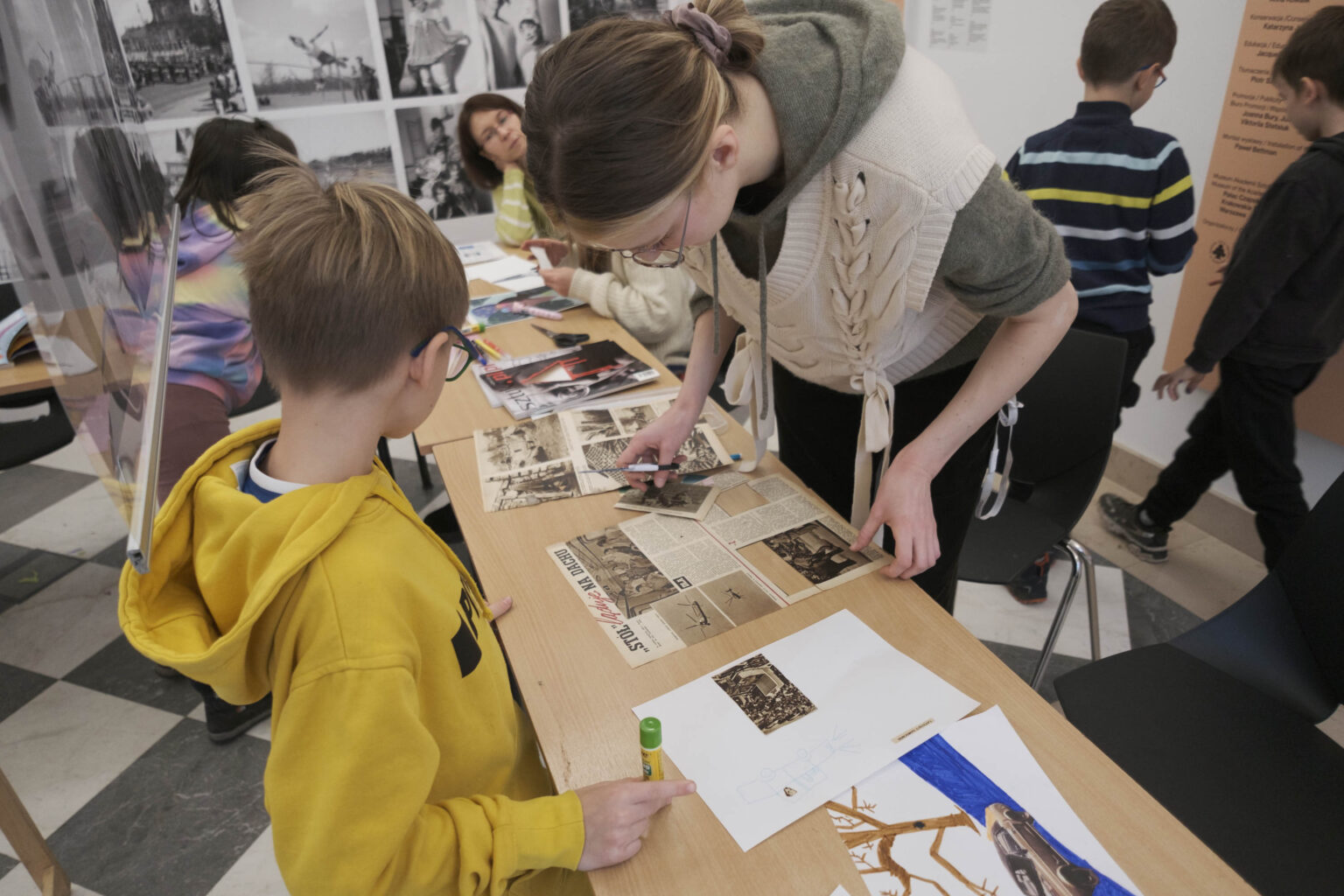 chłopiec w żółtej bluzie i młoda kobieta przy stole z gazetami i materiałami plastycznymi