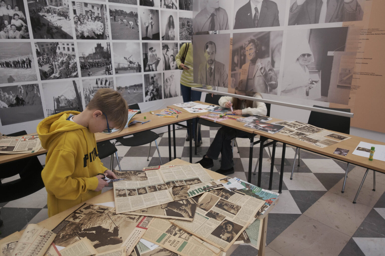 chłopiec w żółtej bluzie przy stole pełnym starych gazet i magazynów, wokół wyeksponowane zdjęcia