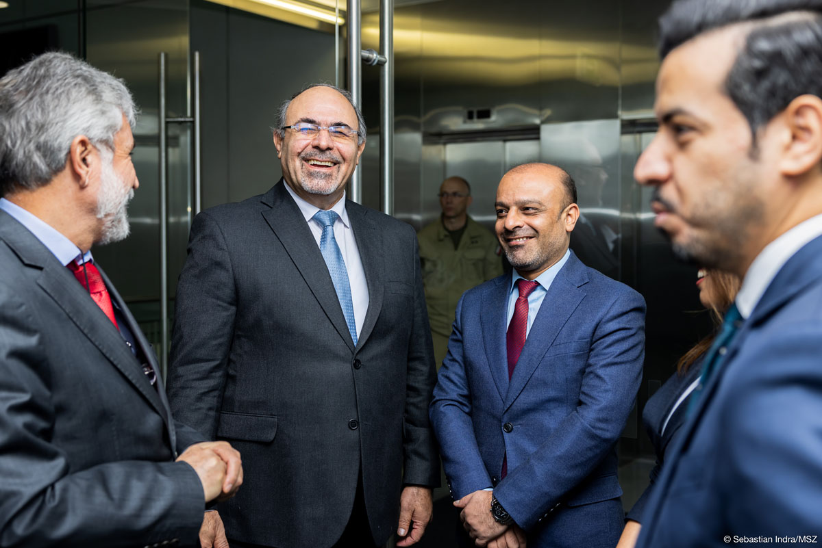 Czterech mężczyzn ubranych w garnitury i krawaty stoi w holu przy windach, uśmiechają się.