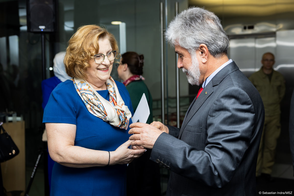 Kobieta w granatowej sukience i apaszce w okularach oraz mężczyzna w szarym garniturze z siwymi włosami i brodą przekazują coś sobie do rąk, uśmiechają się. W tle hol z windami.