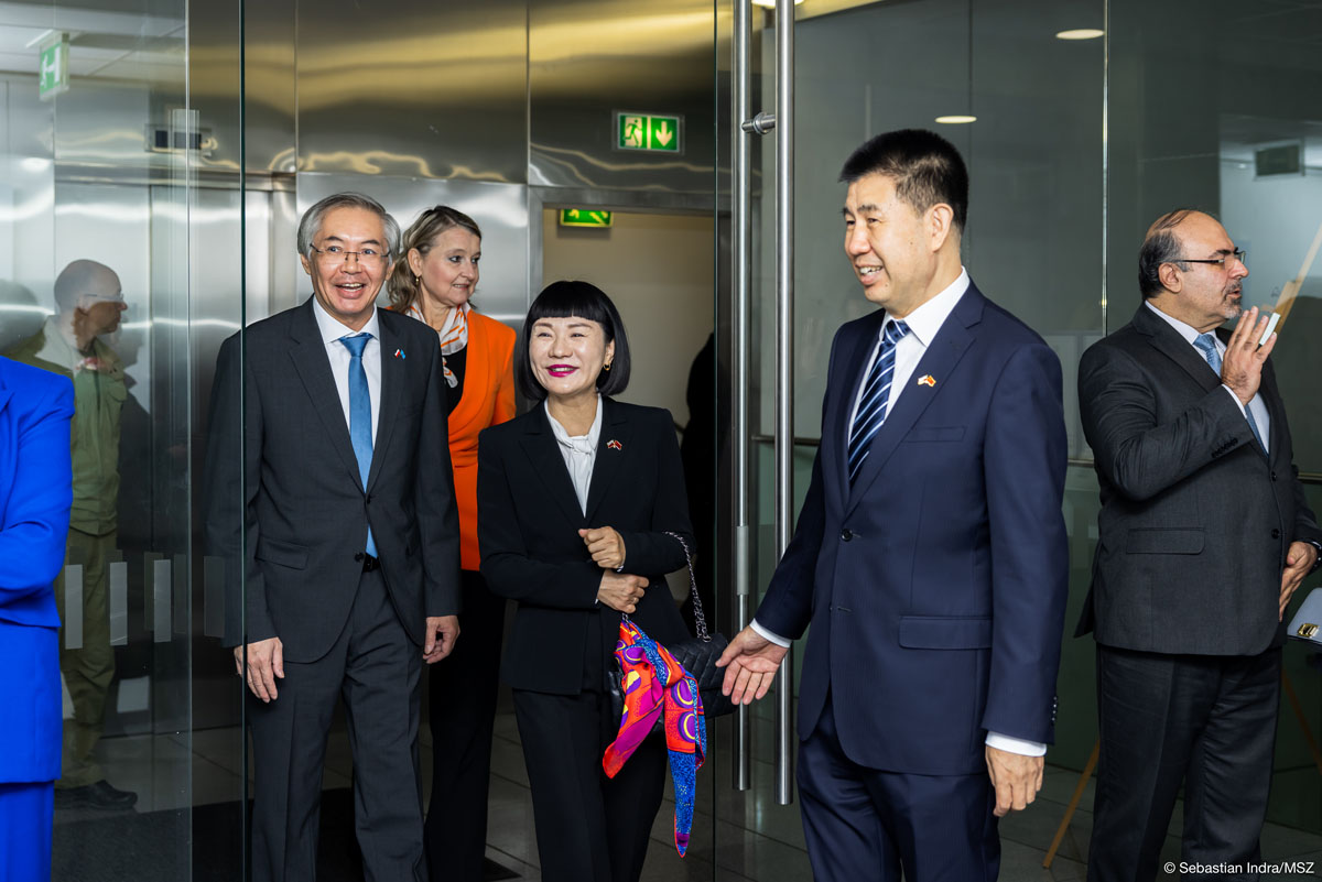 Grupa elegancko ubranych ludzi – kobiet w marynarkach z apaszkami i mężczyzn w garniturach - wchodzi przez szklane drzwi do pomieszczenia. Uśmiechają się.