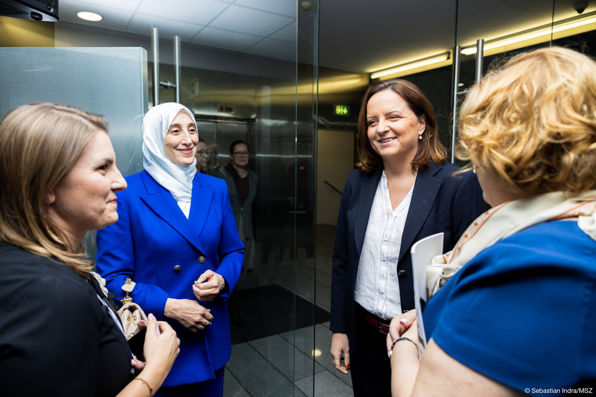 Cztery kobiety stoją w okręgu, rozmawiają ze sobą uśmiechając się – są ubrane w marynarki lub sukienki w odcieniach granatu, trzy mają odsłonięte półdługie włosy, jedna z nich ma na sobie biały hidżab.