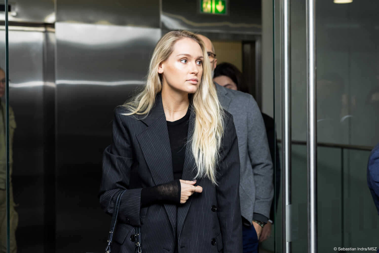 Kobieta z długimi blond włosami w czarnej marynarce, z torebką przewieszoną przez prawe przedramie wychodzi z widny, zerka w lewą stronę. Za nią dwie osoby.
