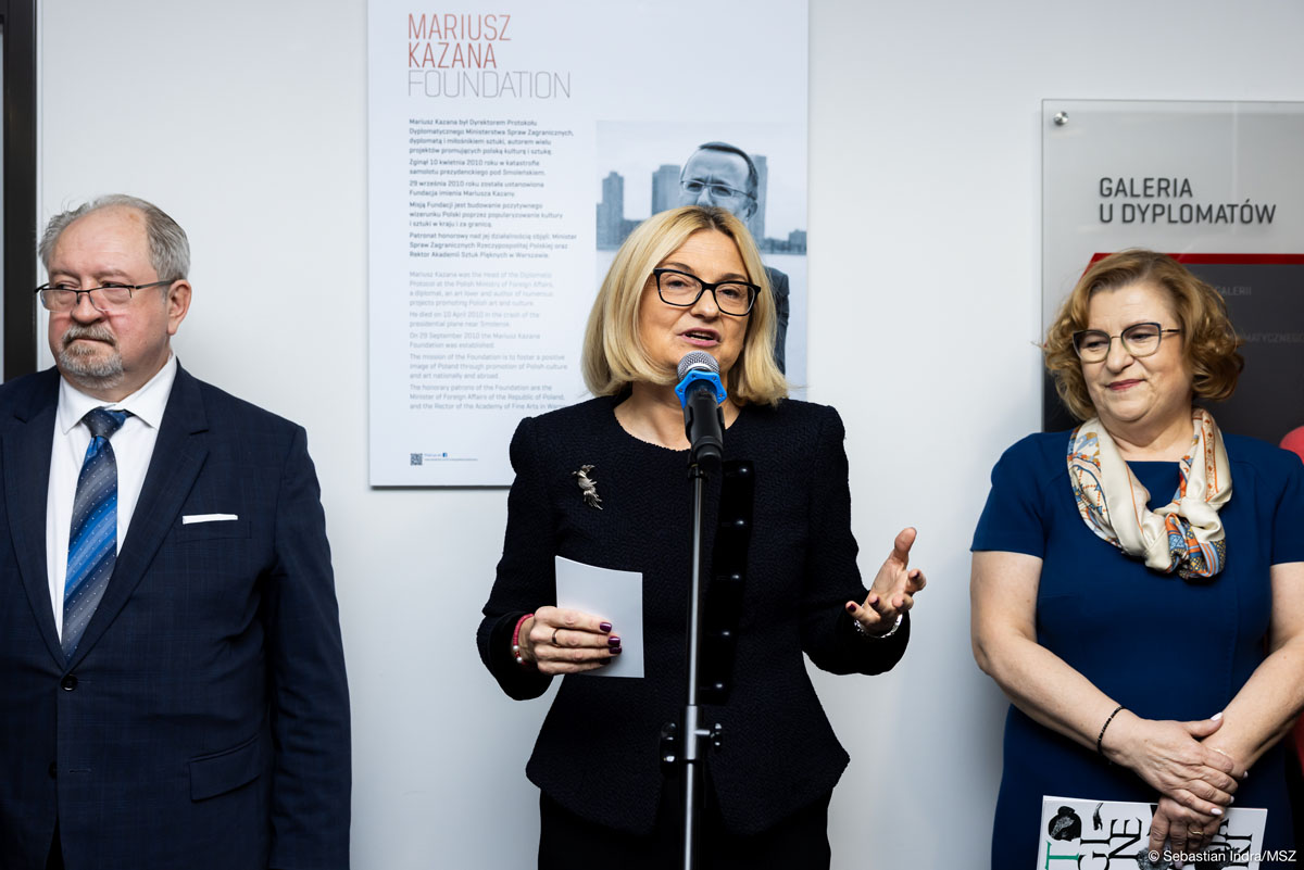Grupa trzech osób stoi za mikrofonem. Kobieta z blond półdługimi włosami w okularach przemawia gestykulując, po lewej stoi mężczyzna w garniturze, po prawej kobieta w granatowej sukience z apaszką. W tle znajduje się tablica z napisem "Mariusz Kazana Foundation" oraz oznaczenie "Galeria u Dyplomatów".