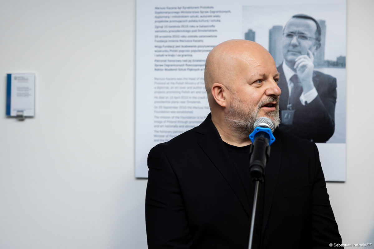 Mężczyzna z brodą, ubrany w czarną marynarkę, przemawiający do mikrofonu. W tle znajduje się tablica informacyjna oraz czarno-biała fotografia mężczyzny w garniturze, trzymającego rękę przy twarzy.