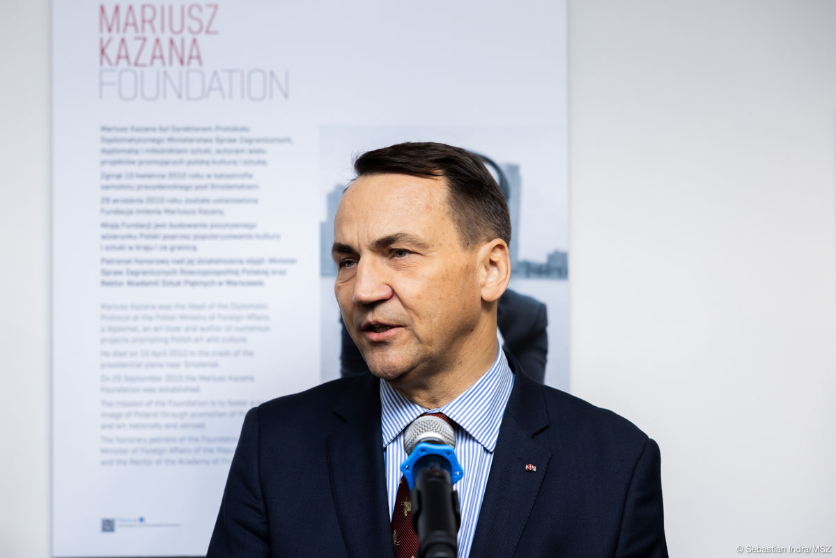 Mężczyzna w granatowej marynarce i koszuli w biało-niebieskie paski przemawia do mikrofonu. Za nimi widać - tablicę z napisem Mariusz Kazana Foundation