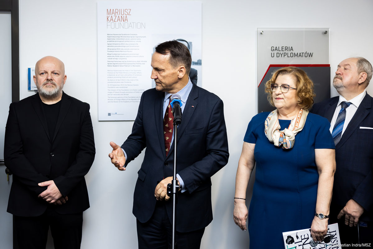 Grupa czterech osób stoi za mikrofonem. Gestykulując, przemawia mężczyzna w garniturze, a pozostałe osoby stoją obok niego, uważnie słuchając. W tle znajduje się tablica z napisem "Mariusz Kazana Foundation" oraz oznaczenie "Galeria u Dyplomatów".