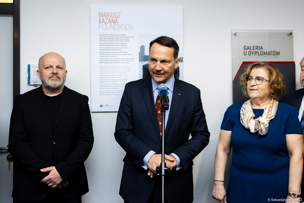 Trójka osób, w tym mężczyzna przy mikrofonie w garniturze, przemawiający do zgromadzonych. Po jego lewej stronie stoi mężczyzna w czarnej marynarce, a po prawej kobieta w niebieskiej sukience i apaszką. Za nimi widać tablicę z napisem "Mariusz Kazana Foundation".