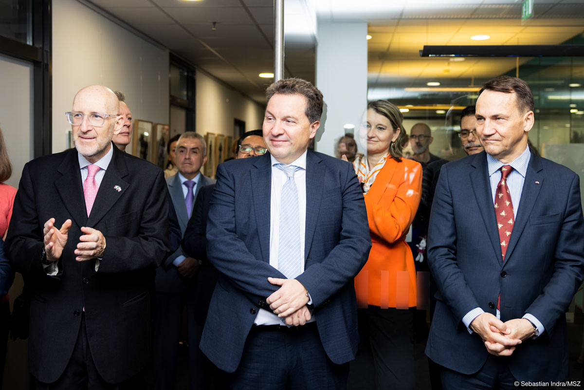 Grupa kilkunastu elegancko ubranych osób patrzy w lewą stronę lekko się uśmiechając. W tle na białych ścianach widać rząd grafik oprawionych w drewniane ramki.