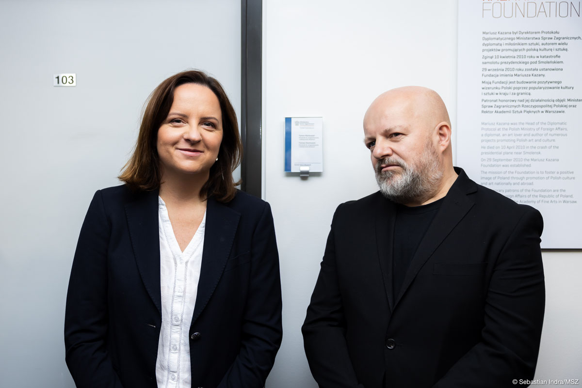 Po lewej kobieta z ciemnymi półdługimi włosami w białej bluzce i czarnej marynarce oraz po prawej łysy mężczyzna z siwą brodą w czarnej bluzce i marynarce stoją na tle białej ściany. Na ścianie wisi plansza z napisem „Foundation” i tekstem zapisanym drobną czcionką. Kobieta patrzy do przodu lekko się uśmiecha, mężczyzna patrzy w bok poważny, zamyślony.