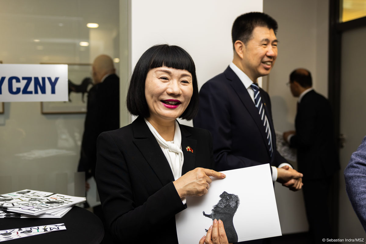 Kobieta z półdługimi czarnymi włosami i krótką grzywką elegancko ubrana w czarną marynarkę i białą koszulę uśmiecha się do obiektywu, w rękach trzyma katalog. W tle ciemnowłosy mężczyzna w garniturze z krawatem uśmiecha się, dalej inni ludzie oglądający grafiki na ścianach, na szklanej szybie fragment napisu: „CZNY”.