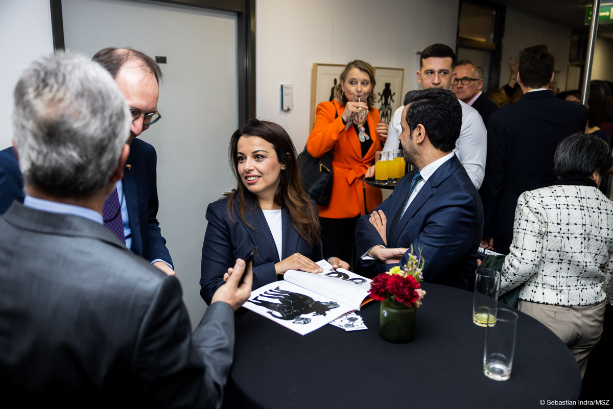 Kilkoro elegancko ubranych ludzi stoi przy wysokim stoliku barowym pokrytym czarnym obrusem, na którym stoją dwie szklanki i czerwono-żółte kwiaty w niskim czarnym wazonie. Kobieta trzyma w rękach otwarty katalog z czarno-białymi grafikami, patrzy w stronę dwóch mężczyzn. W tle kelner z tacą z żółtymi napojami w szklankach, za nim kobieta w pomarańczowym żakiecie pije ze szklanki, dalej inni ludzie.